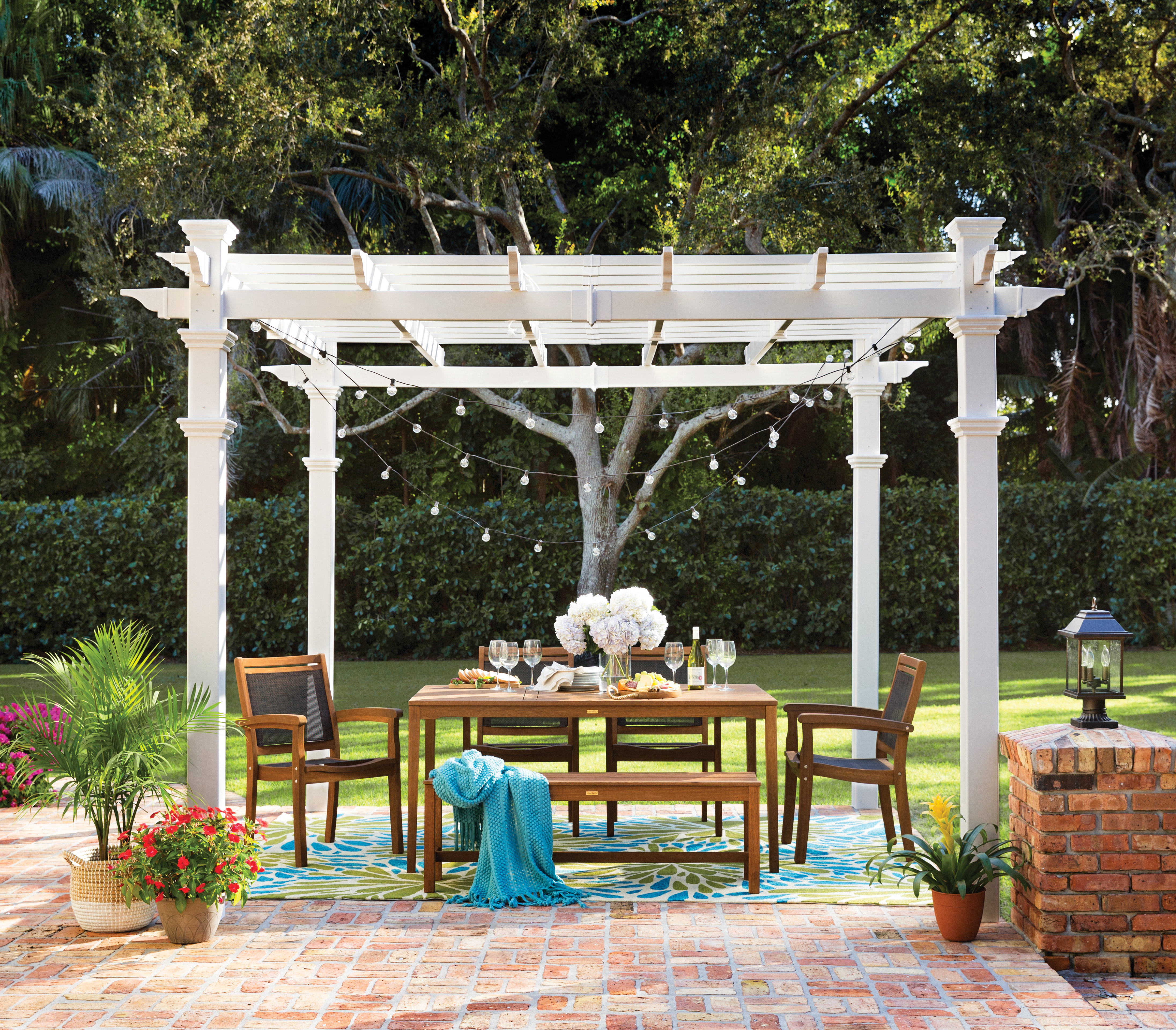 brick patio dinner set shaded by a white pergola decorated with string lights